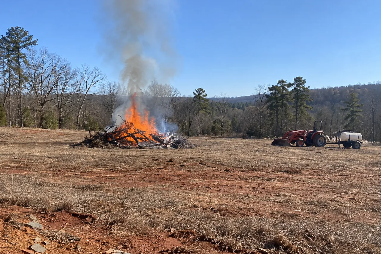 Brush pile burning in the middle of a wide cleared field, with a tractor and water tank parked well back