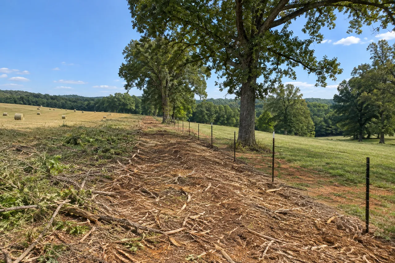 Freshly mulched strip along a barbed-wire fence between a hay field and a pasture, with big oaks left standing